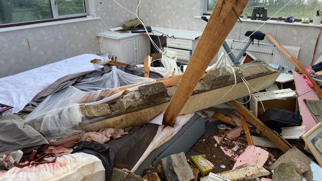 A bedroom full of stone debris and broken furniture and wooden beams. There is a bed with a mattress with a window lintel across it and a roof timber penetrating the mattress.