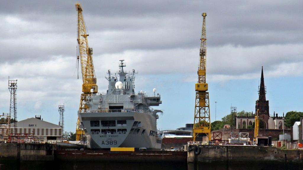 A ship is in the Cammell Laird shipyard. There are two yellow cranes and Birkenhead Priory is to the right.