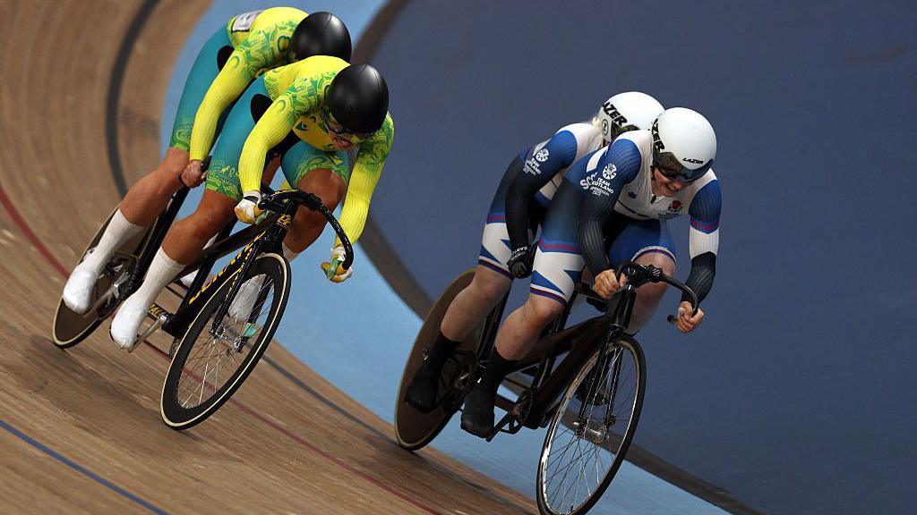 Two pairs of tandem cyclists racing against each other on a cycling track. One team are wearing light yellow and green. The other team are wearing blue and white.