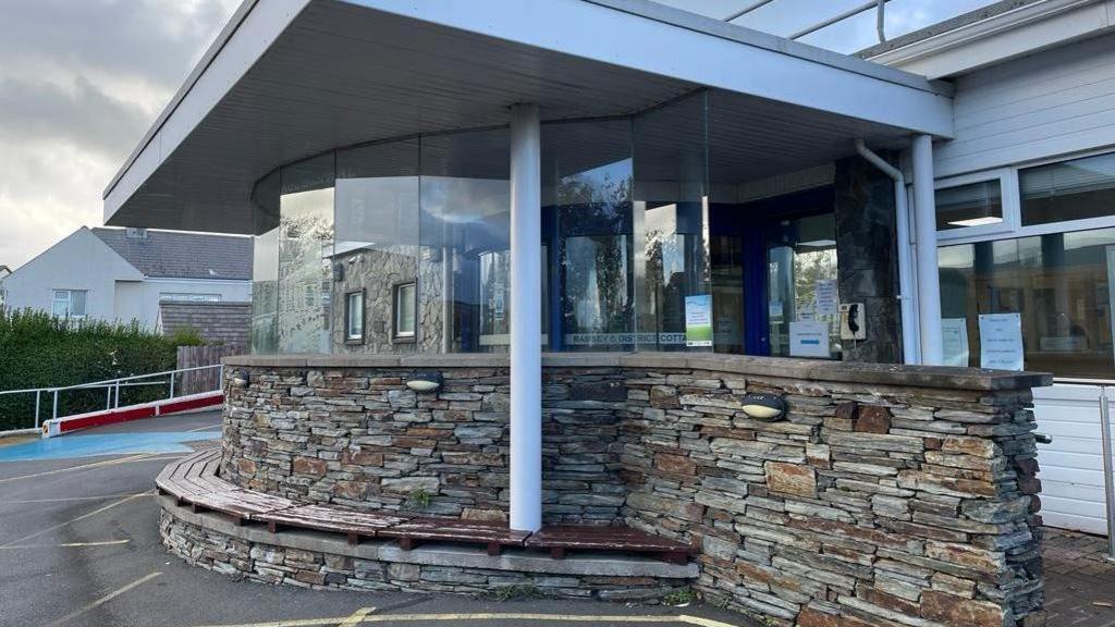 The entrance to Ramsey Cottage Hospital, which is a blue revolving door under a white canopy surrounded by a stone wall.