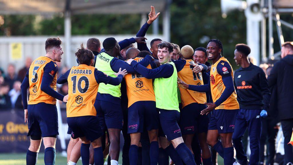 Slough Town players wearing orange shirts and blue shorts celebrating, including with substitutes, after scoring against Grimsby at Arbour Park in November 2023