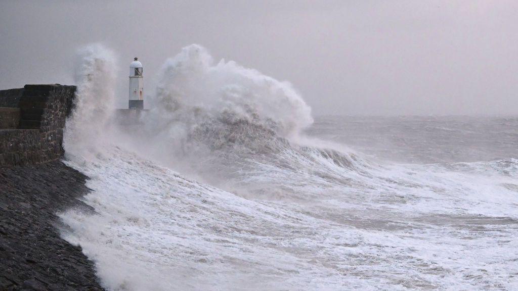 A big wave rushes towards a light house on a grey stormy day.