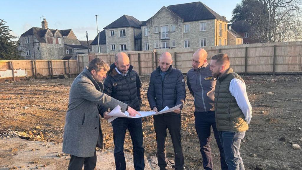 A group of men stand at a levelled building site which is surrounded by a wooden fence and homes. They are all in coats looking at a large piece of paper which looks like a plan.