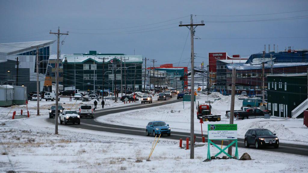 A road in Iqaluit that shows several buildings, various cars on the road, lots of power poles and somewhat light snow on the ground.