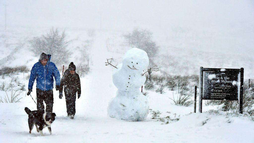 A father and son walking their black and white dog on Black Mountain in the snow of December 2017. There is a large snowman next to them.