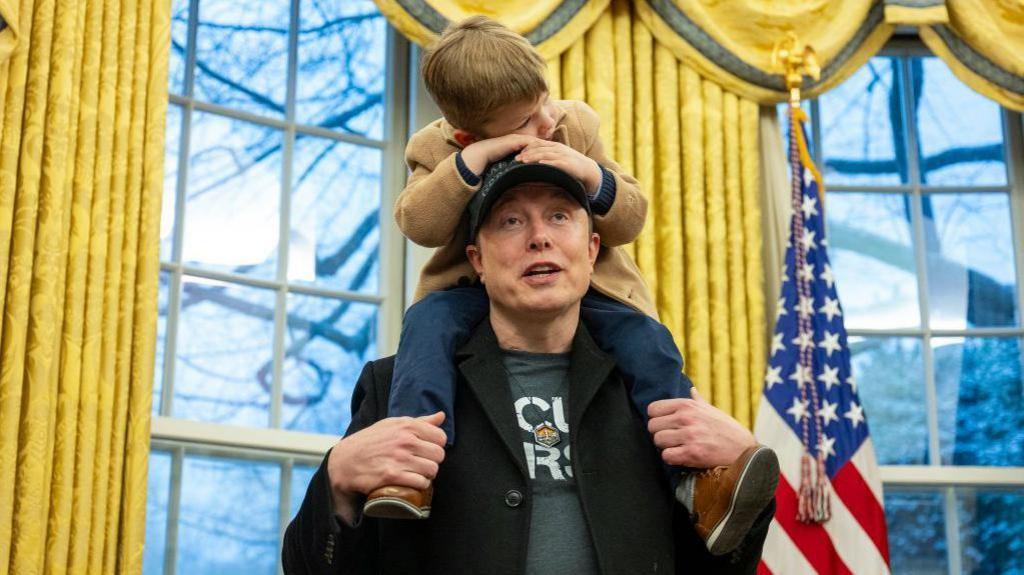 Elon Musk stands in the Oval Office of the White House with his young son sitting on his shoulders with his hands on his father's head. Windows with golden curtains and the US flag can be seen behind them.