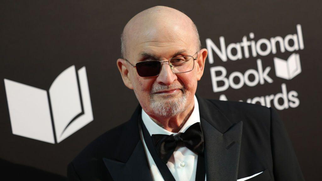 Sir Salman Rushdie pictured wearing a tuxedo and a black bow tie. He is wearing glasses, with one side darkened after he was blinded.