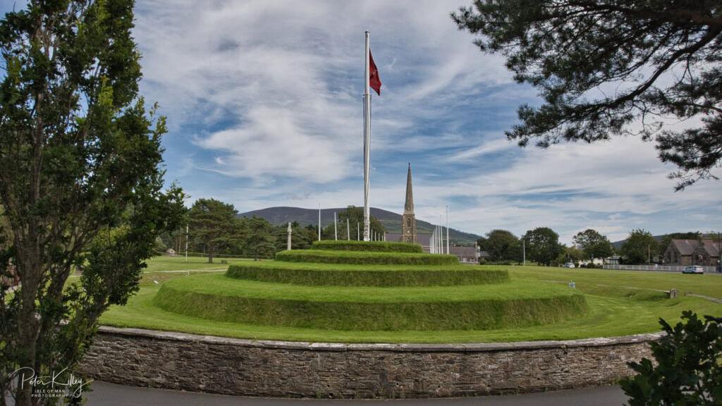 Tynwald Hill can be seen with five grassed tiers surrounded by a stone wall at the bottom with a flag pole carrying a Manx flag in the centre. The spire of the Royal Chapel of St John's can be seen in the distance.