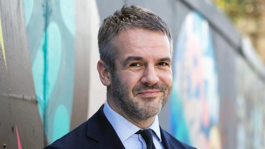 A man with short dark hair and stubble is wearing a dark navy suit, blue shirt and dark tie. He is facing the camera with a slight smile