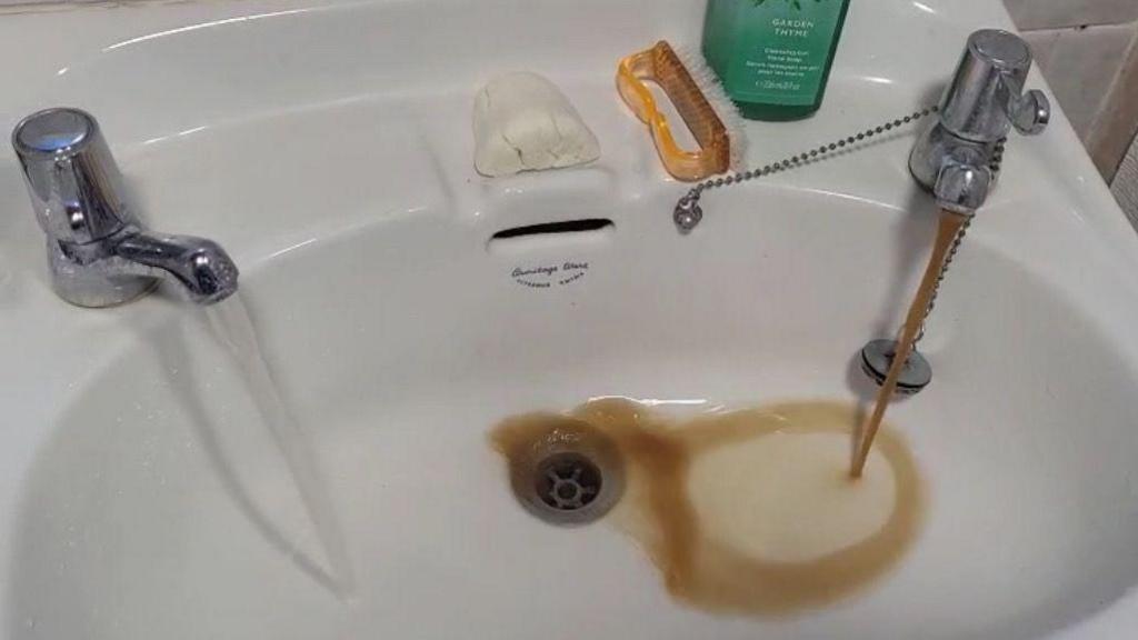 Brown water coming out the right tap in a sink. The left tap is running clear water. The sink is white and there is a bar of soap, an orange nail cleaner and soap on the side.