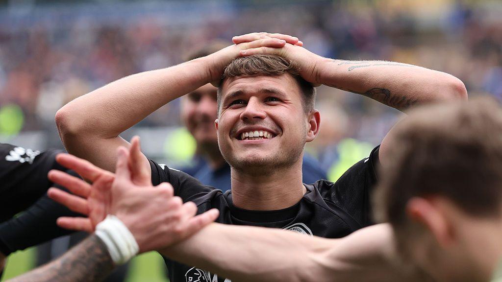 Boreham Wood's Zak Brunt, wearing a black football shirt, stands among team-mates with his hands on his head while smiling