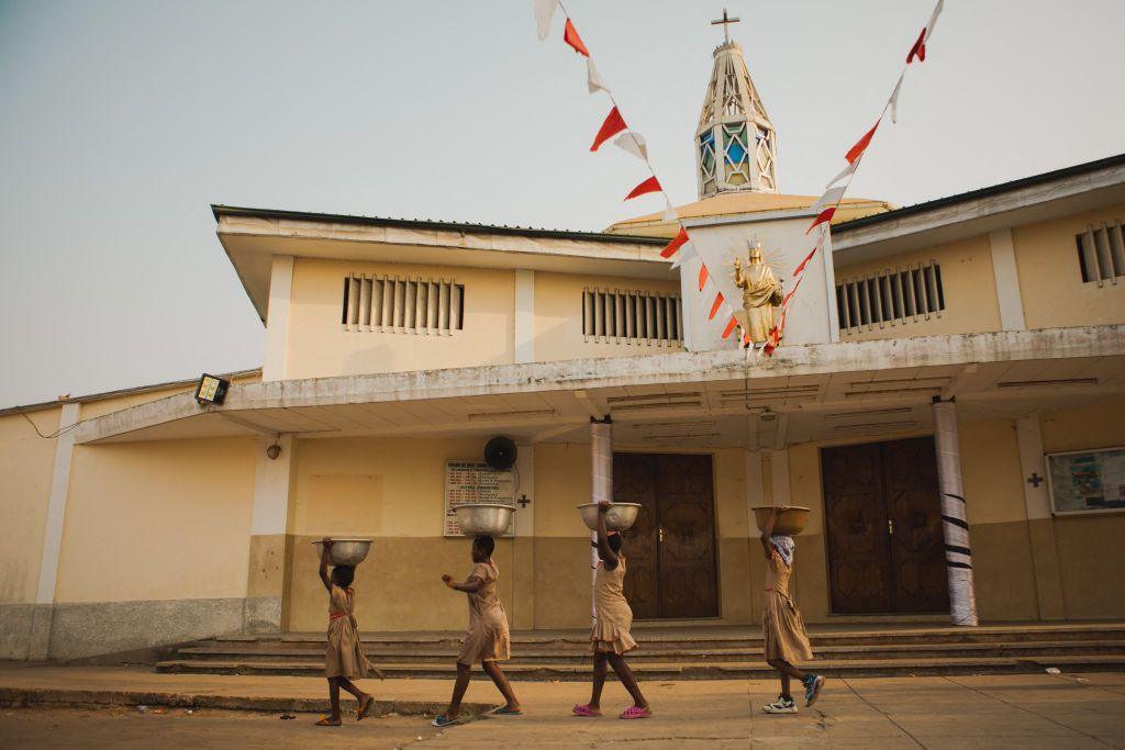 Four girls walk in line past a church. Each carries a large bowl of water on her head. 