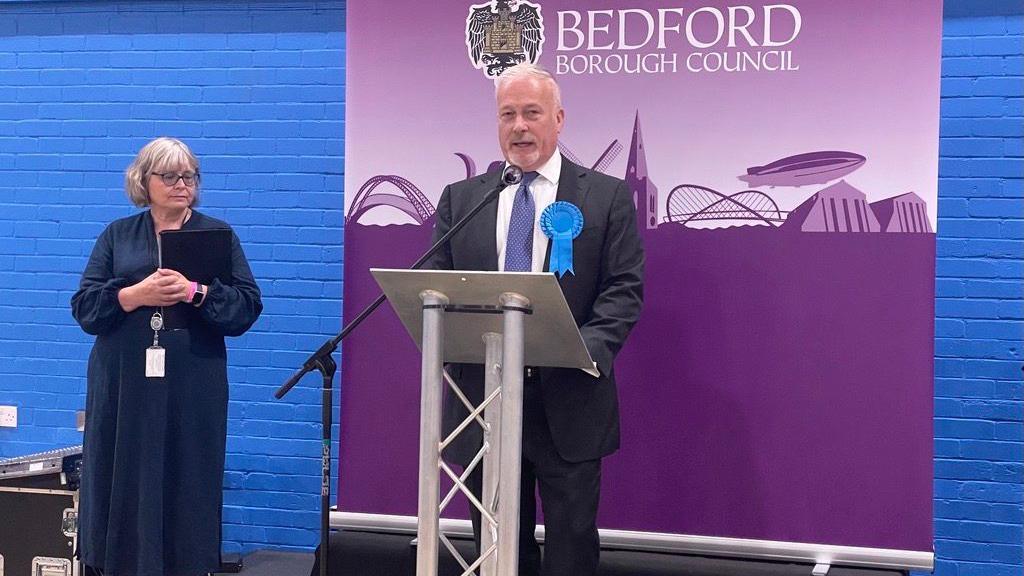 Richard Fuller on stage and speaking from a podium at the count, with a woman standing to his right. He is wearing a dark suit with a blue rosette attached to his lapel. 