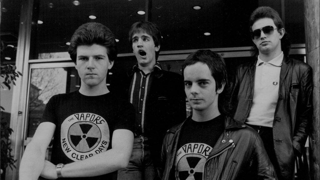Four young men, two of whom are wearing The Vapors t-shirts. One man is yawning. The image is in black and white.
