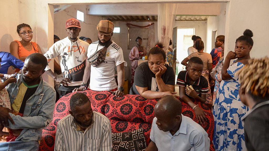 A family group look mournful as they stand behind a coffin wrapped in a red patterned cloth.