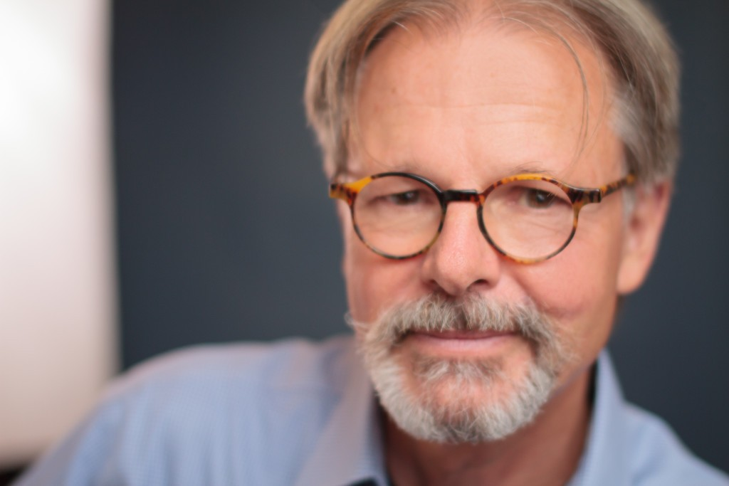 A man in a shirt with glasses and a well-kemp grey beard smiles.