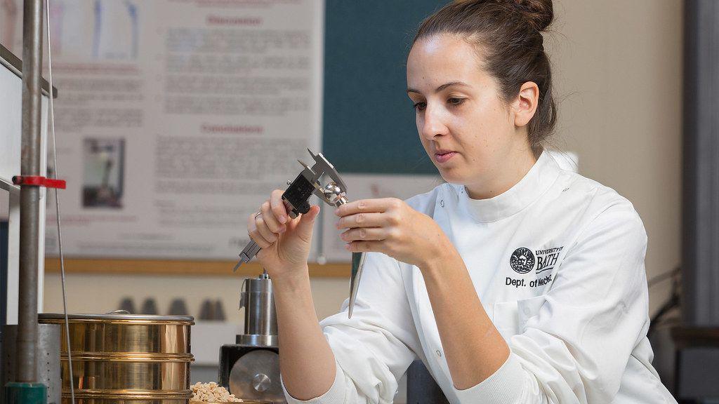 A woman in a white lab coat is measuring with a device in her hands. She is in a laboratory and there are research posters on the wall behind her.