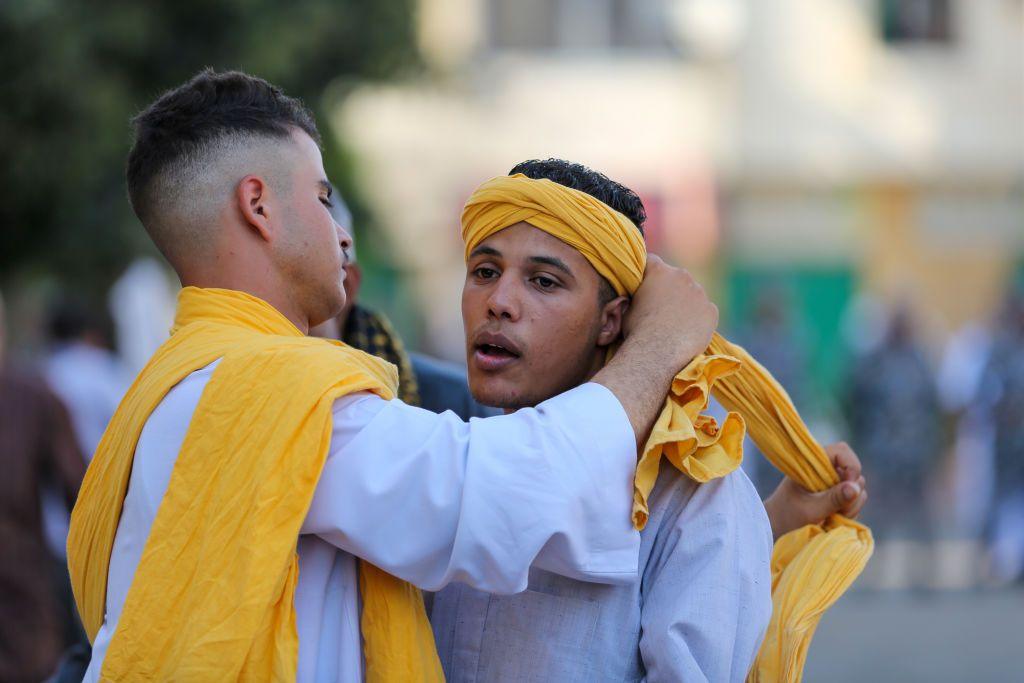 Men in matching white robes wrap yellow fabric around one another.