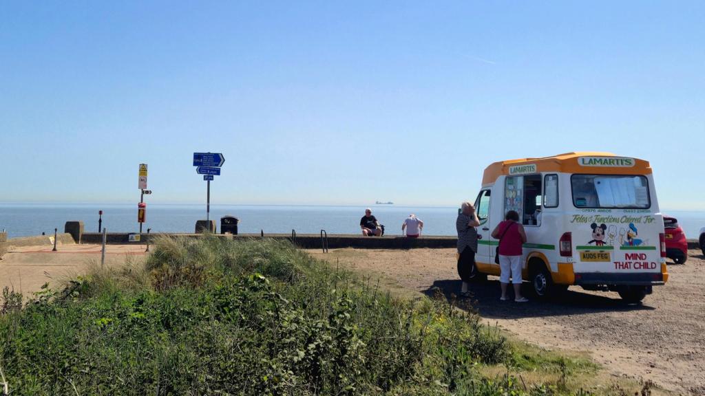 Two people stand by ice cream van as they wait for their ice creams, with the sea in the background