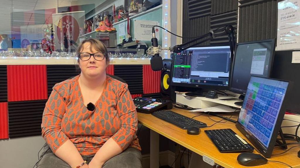 Georgy Tubb has fair hair tied-back from her face and wears glasses. She is wearing an orange and grey shirt sits and grey trousers and sitting behind a desk with keyboards and computer screens in a radio station