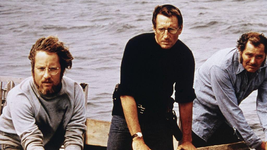 Three men in a boat. Richard Dreyfuss in grey sweat shirt and blue jeans, Roy Scheider in black jumper and jeans, Robert Shaw in blue work shirt and jeans. They are standing in a boat at sea. 