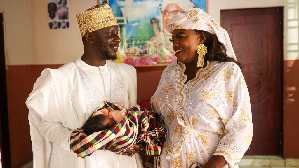 A couple - wearing white and gold outfits - gaze at each other white holding a newborn baby in Banjul, The Gambia - 15 February 2025.