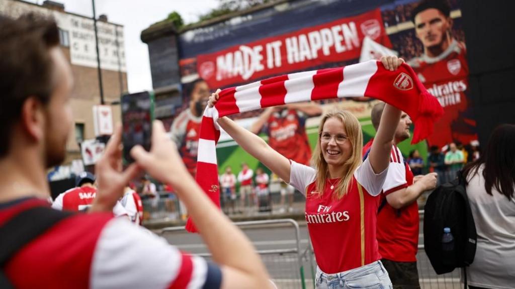 A woman holds up her Arsenal scarf as someone takes a picture of her - the words 'made it happen' are written on a banner in the background