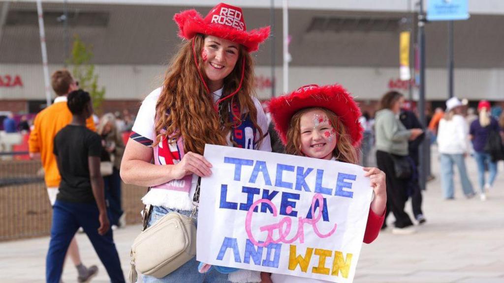 A young woman and a girl, both with long red hair and sporting red cowboy hats emblazoned Red Roses. They are smiling and holding a placard reading "Tackle like a girl and win".