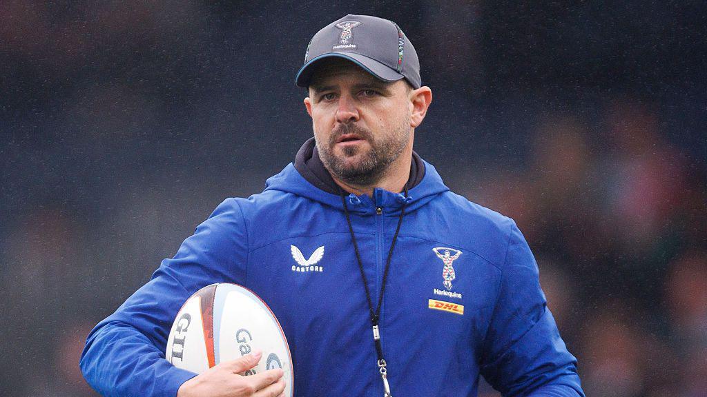 Nick David stands wearing a black cap, a blue jacket and holds a rugby ball to his chest under his right arm