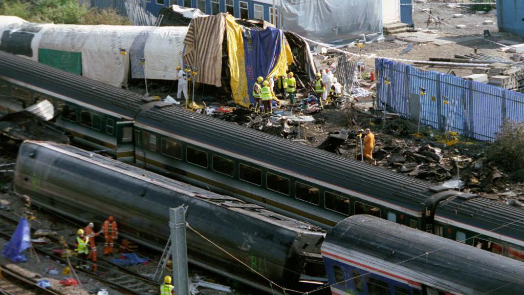 The debris from the crash with rail workers in high vis jackets and hard hats on the track