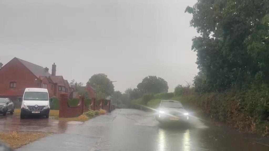 A flooded road with a car driving slowly through the water. There are houses on the left, hedge on the right.