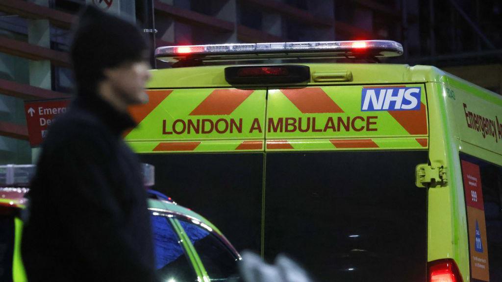 A London Ambulance Service vehicle is seen parked outside an emergency department at night. The NHS logo is displayed prominently on the vehicle. A silhouette of a person walking in the foreground is slightly out of focus.