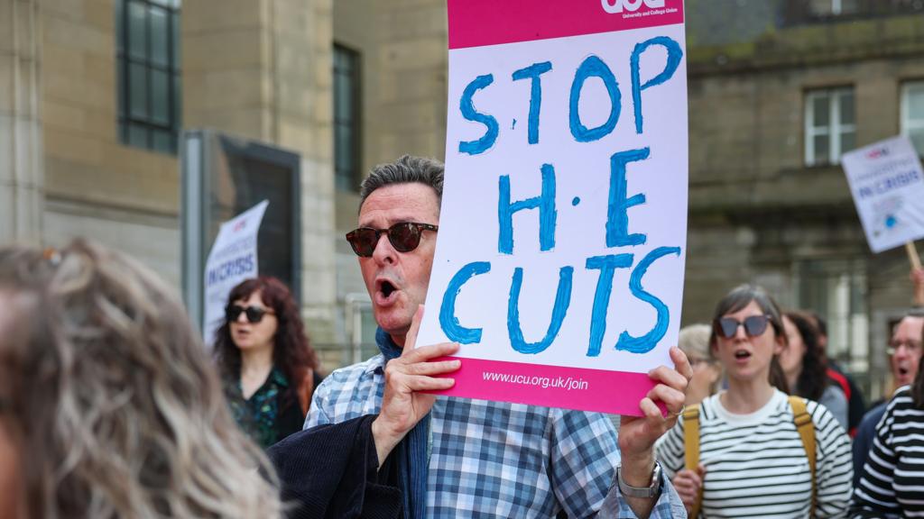 A man in sunglasses and a checked shirt holds a sign which says: Stop the Cuts