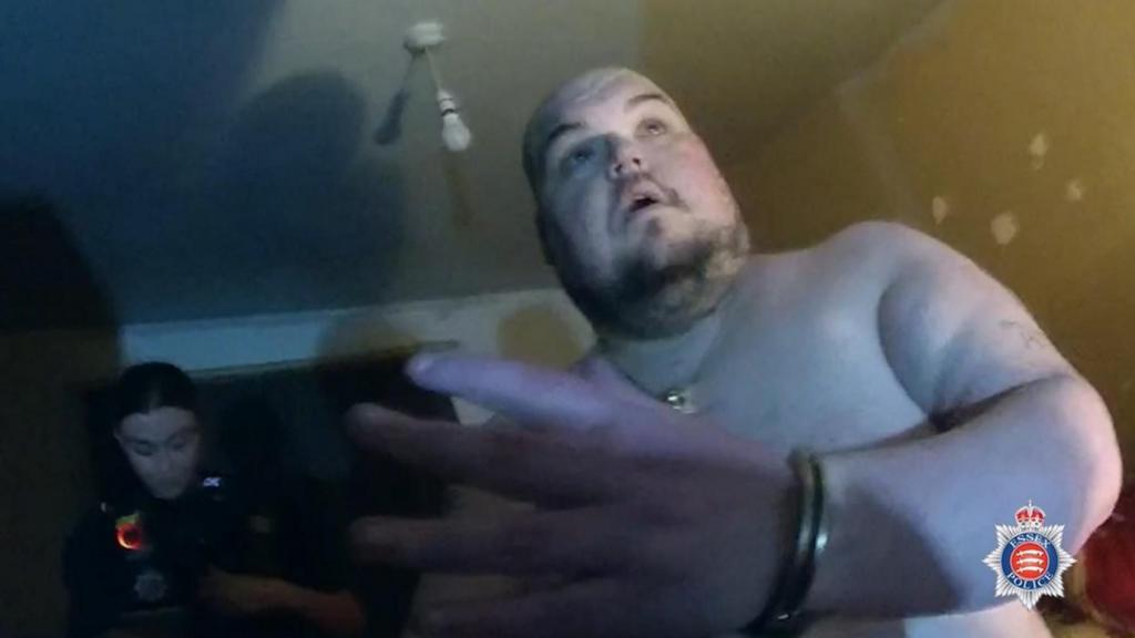 Gavin Plumb being arrested inside his house. He is topless. A woman police officer is standing in a doorway behind him.