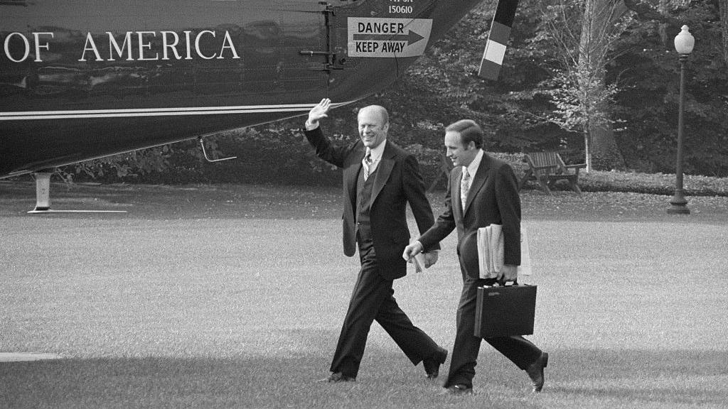 President Gerald Ford waves as he walks past the presidential helicopter with Dick Cheney walking next to him, shown in 1975 in a black and white photo.