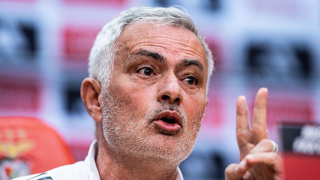 Close-up of Jose Mourinho speaking at a press conference. He has short grey hair, brushed to his right, and stubble. He is gesturing with his left hand.