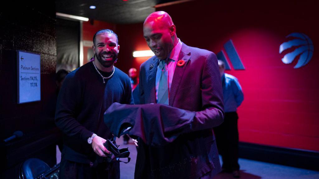 Two men stand in a darkened hallway inside a stadium. One looks into the camera, laughing. The other, dressed in a suit and tie, is holding a basketball jersey in his hands and looking down at it, smiling.