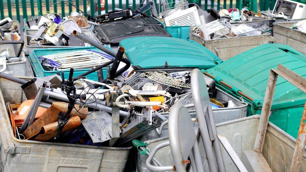 Eight large bins with green lids, full to the brim and overflowing with scrap metal. Discarded items include clothes hangers, bike frames, metal chairs, ironing boards, microwaves and poles.