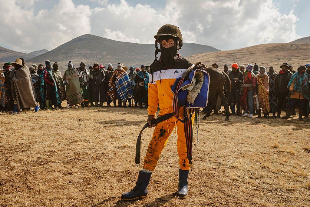 A young jockey poses for a portrait. People stand in the background behind him, and the mountains rise in the distance.