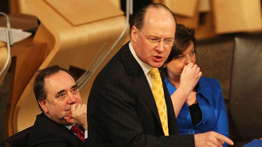 John Swinney speaking in the Scottish Parliament while finance minister with Alex Samond behind him