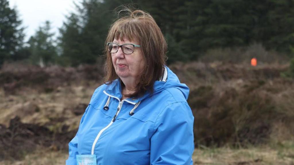 Dympna Kerr stands with a blue raincoat on in the middle of boglands in County Monaghan. Ms Kerr is an older woman with shoulder-length brown hair and a full fringe. She is wearing a pair of tortoise shell glasses with turquoise legs and is looking away from the camera.