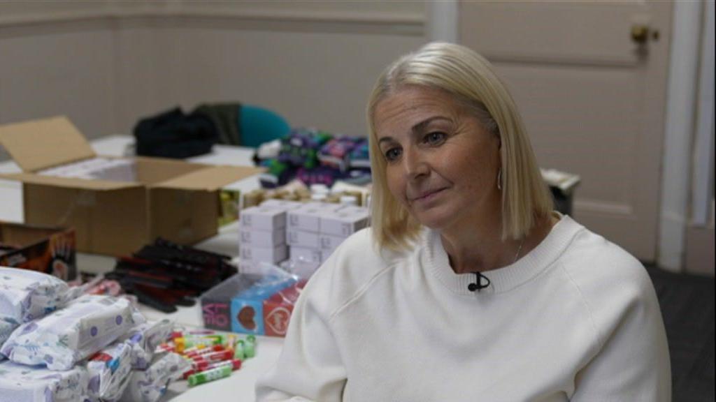 Vicky Graham has a platinum blonde bob and is wearing a white jumper. She is sitting in front of a table with items across it, including wipes and hand warmers.