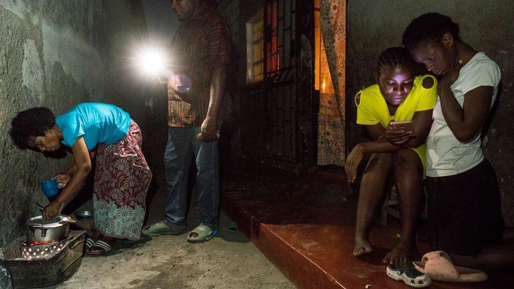 A family scene showing people dealing with power cuts. On the left, a woman is cooking by the light of a mobile phone and in the right, two people are looking at a phone.