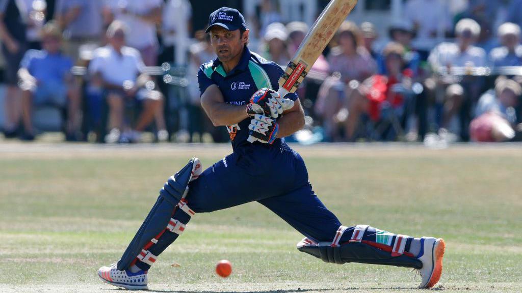 Cricket player Mark Ramprakash in full gear, including pads, gloves, and a helmet, is captured mid-action playing a shot. He is wearing a dark blue uniform and is positioned in a low stance, extending the front leg forward while swinging the bat toward an orange cricket ball on the ground.
