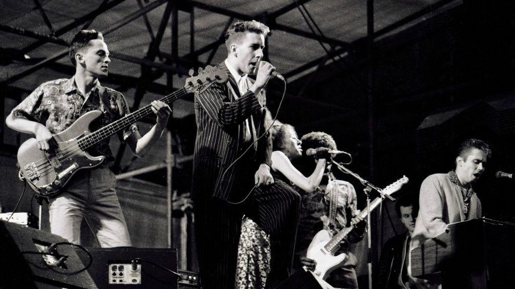 The band the Specials play at a concert at Coventry's Butts Stadium. Bass player Horace Panter is at the far left of the picture next to lead singer Terry Hall who is singing into a microphone. Rhoda Dakar sings into a microphone next to Lynval Golding and Jerry Dammers of the band. 