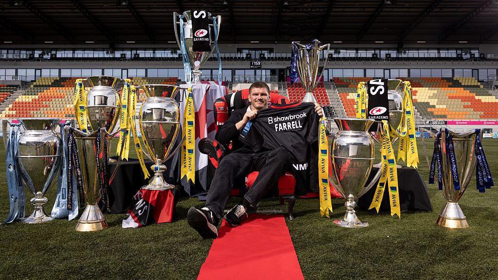 Owen Farrell sits on a red chair in the middle of the StoneX Stadium pitch, holding up a Saracens shirt with his name on the back, and surrounded by trophies he won in his first spell at the club, with a red carpet rolled out in front