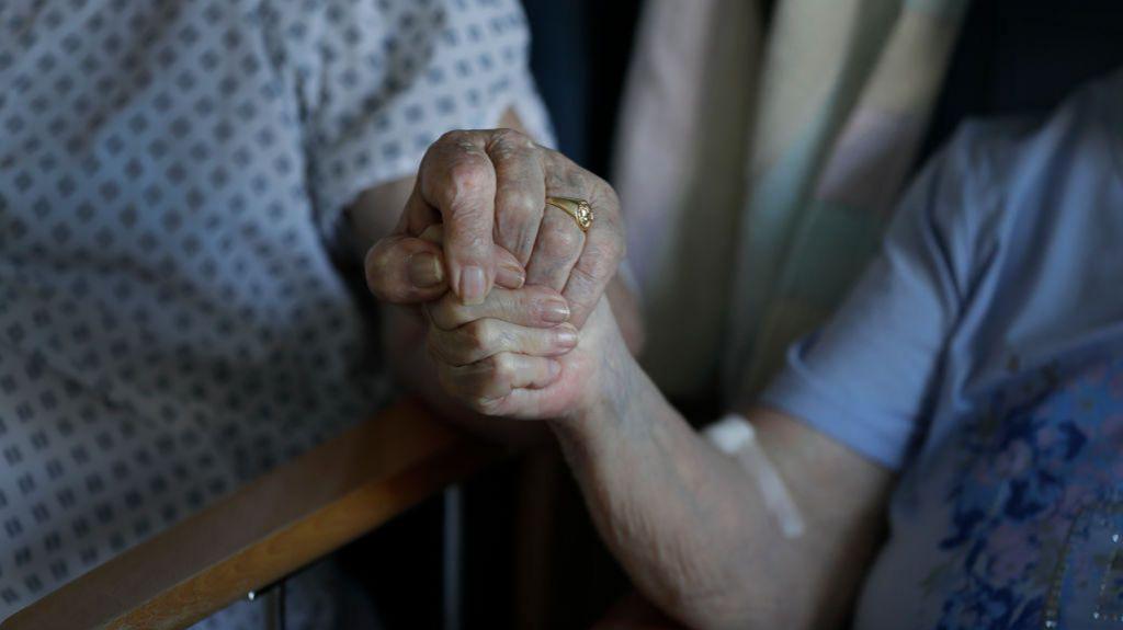 Close up of an elderly couple holding hands. One person wears a hospital gowns and a gold ring. The other is wearing a purple t-shirt. Their faces are not in view. 