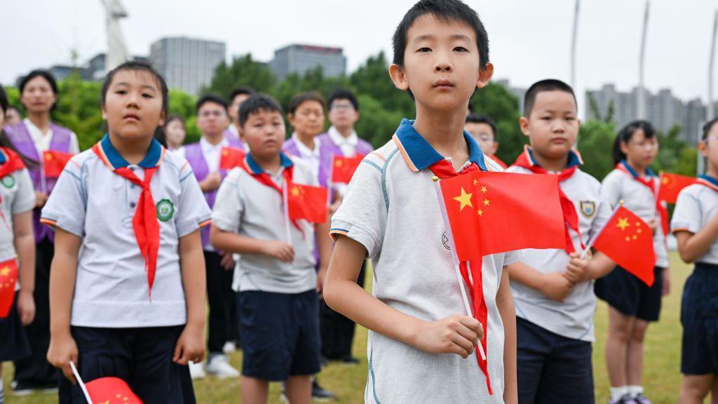 A school boy holds a Chinese flag up in his hand. Behind him are lots of school children in white tops and blue shorts with red and blue and white neckerchiefs. they are standing on a field with tees and high rises in the background