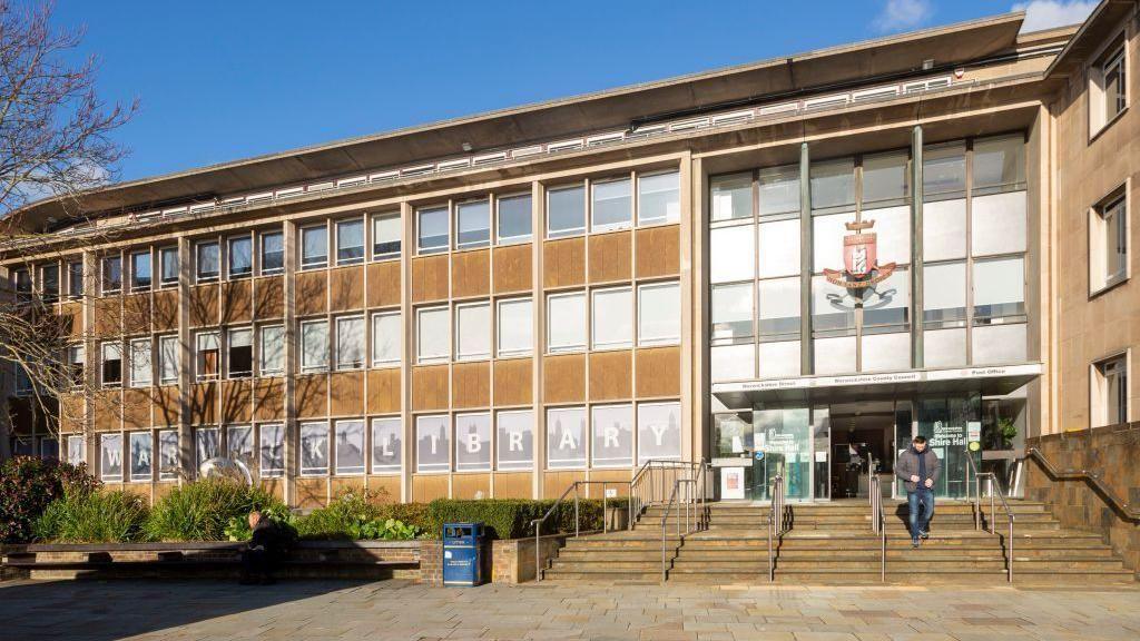 The county council building in Warwick. The building has three floors and lots of windows. The council's logo is on the front of the building. A set of steps leads to the building's glass doors, which are open, with one person walking out of the building down the steps.