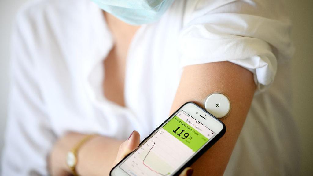 A woman wearing a face mask and a white shirt scanning a blood glucose monitor on her arm using her phone.